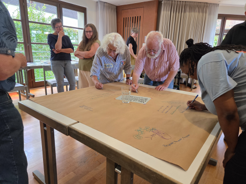 Menschen beugen sich über ein großes Papier auf einem Tisch und schreiben.