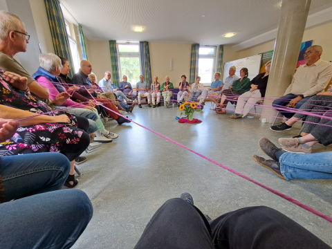 Menschen sitzen in einem großen Stuhlkreis. Jede Person hält einen Teil einer landen Schnur fest. 